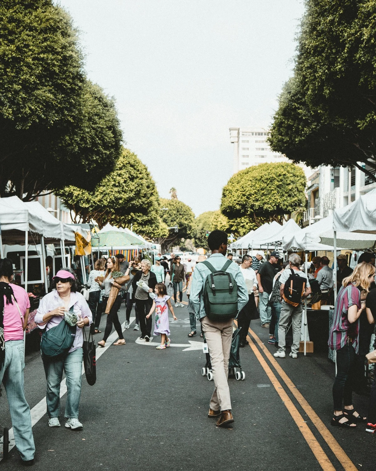 Shoppers browsing vendor stalls at an outdoor farmers market lined with trees